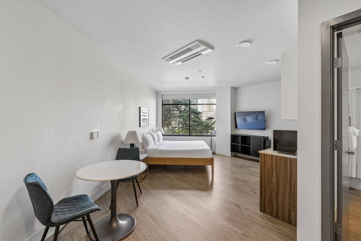 A modern hotel room with a bed, a TV, a dining table with chairs, and a large window.