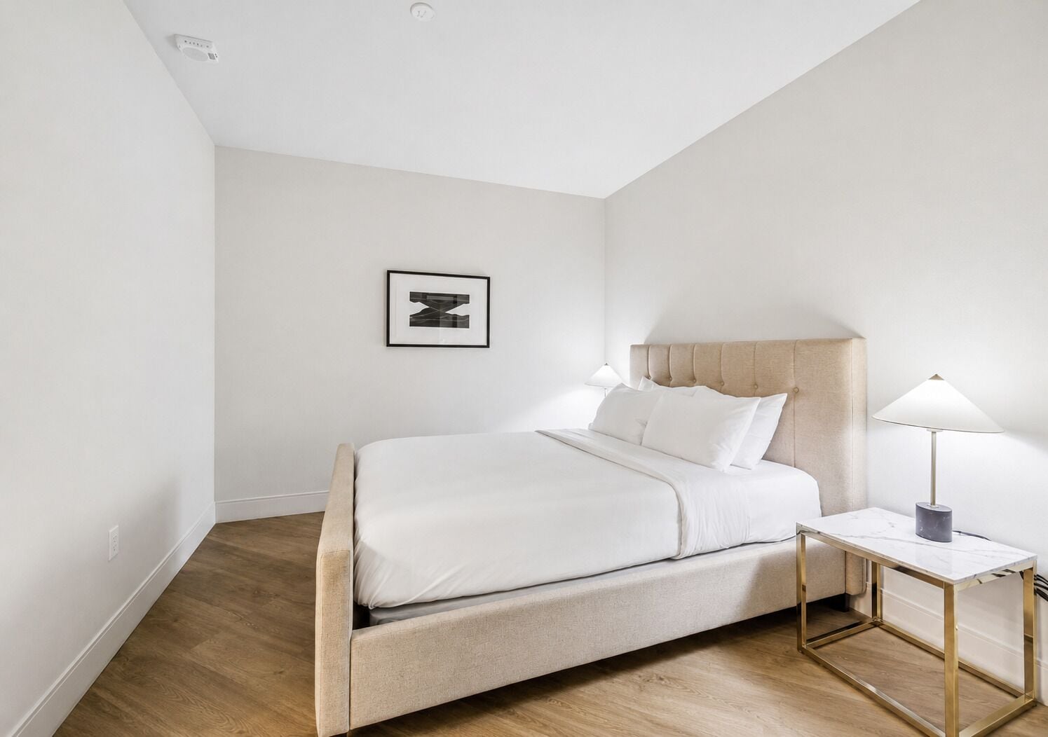 Ein schlicht eingerichtetes Schlafzimmer mit Bett, Nachttischlampen und einem gerahmten Bild an der Wand.