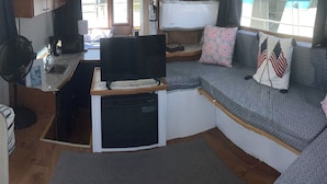Interior compacte d'una autocaravana amb petita cuina, nevera, televisió i zona de seients amb coixins.