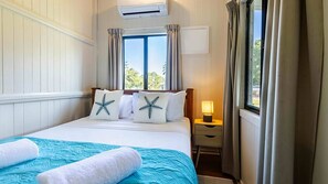 A hotel room with a bed, a bedside table with a lamp, a window with curtains, and an air conditioning unit.