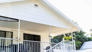 Área externa coberta com guarda-corpo branco, uma cadeira preta e uma janela com cortinas.