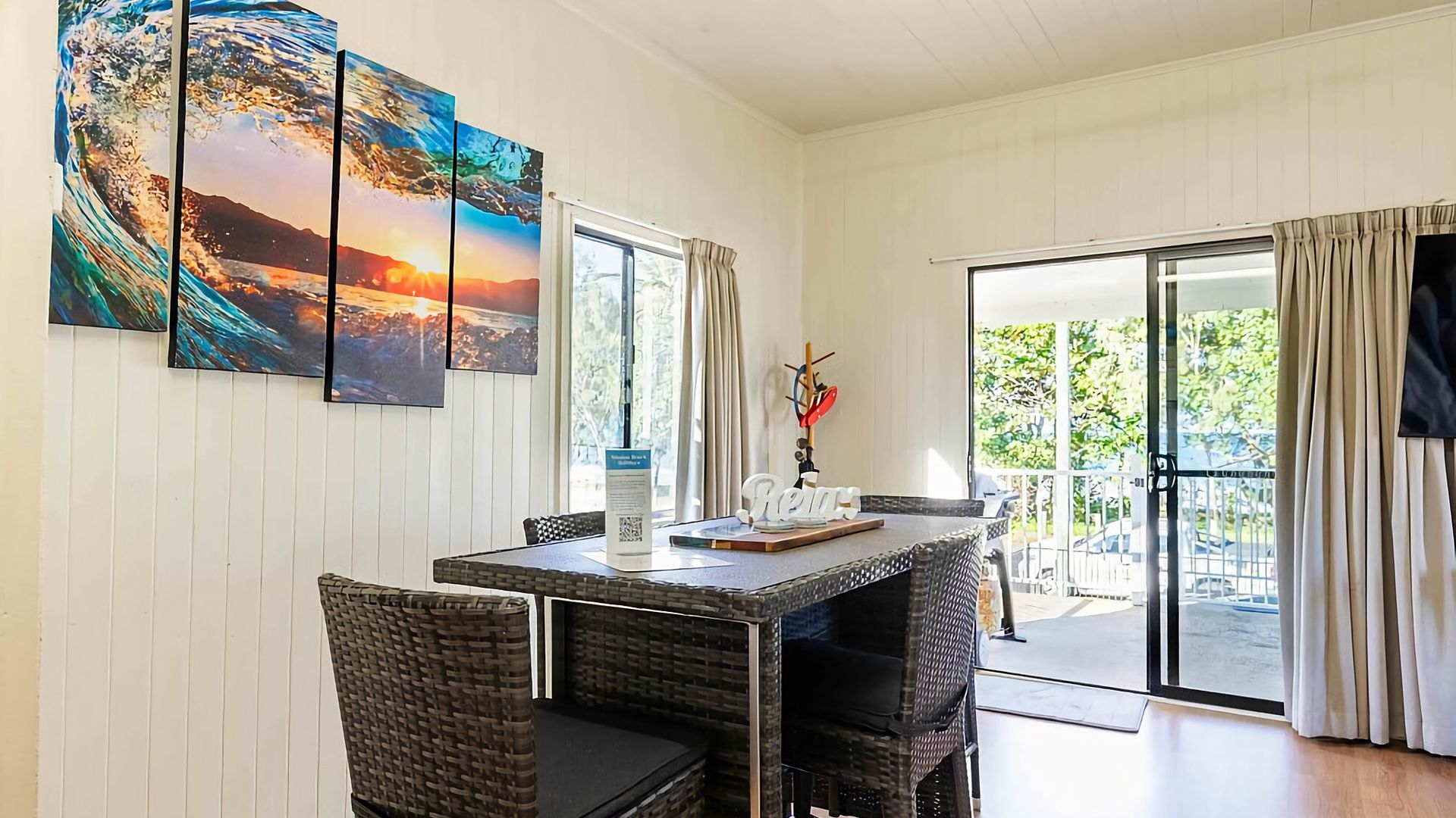 A dining area with a table and chairs, a painting on the wall, and a view through the glass door.