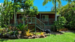 A wooden house with a green porch and a staircase, surrounded by lush greenery and a clear blue sky. - Mission Dreaming - 1 Bedroom Cottage (South Mission Beach)