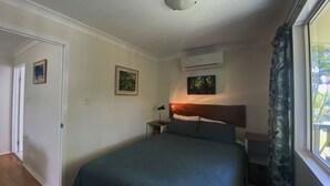 A bedroom with a bed, a nightstand, an air conditioning unit, and a window with curtains. - Mission Dreaming - 1 Bedroom Cottage (South Mission Beach)