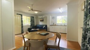 A room with a dining table, chairs, a sofa, a ceiling fan, and a window. - Mission Dreaming - 1 Bedroom Cottage (South Mission Beach)