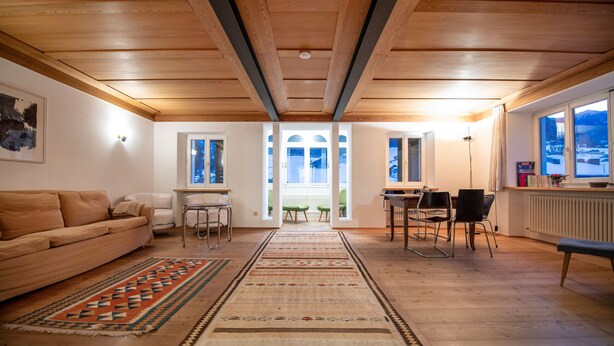 A spacious living area with a wooden ceiling, a sofa, a dining table with chairs, and a view of the outdoors through large windows. - Apartments Stina 122A (Ortisei)