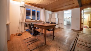 A dining area with a wooden table and chairs, a window with curtains, and a view into the kitchen. - Apartments Stina 122A (Ortisei)