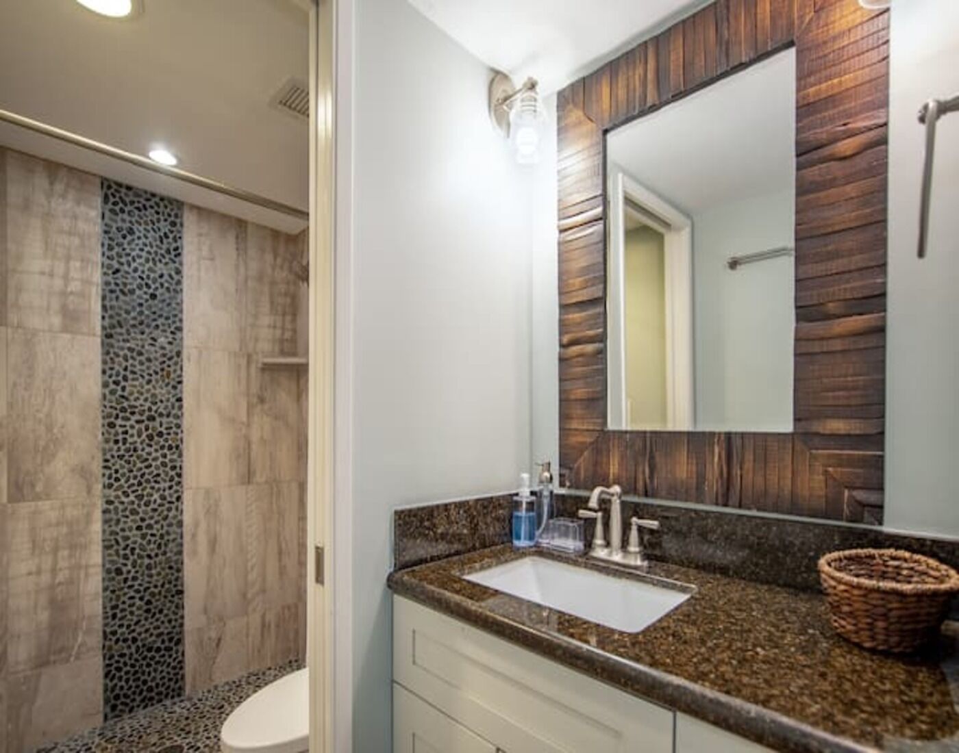 Un bagno con parete in pietra, WC bianco, piano di lavoro in granito, lavabo, specchio e zona doccia.