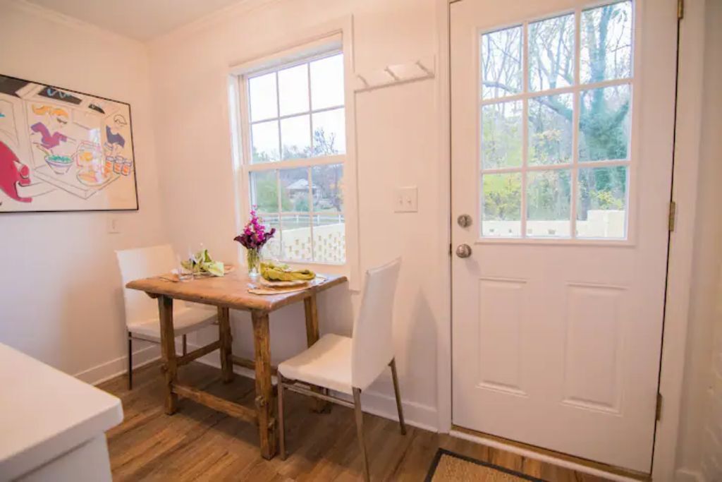 A dining area with a wooden table and white chairs, a vase with flowers, and a large window with a view of trees.