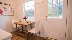 A dining area with a wooden table and white chairs, a vase with flowers, and a large window with a view of trees. - Luxury cottage with pool (Charlottesville)