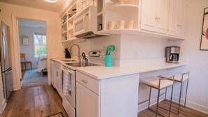 A kitchen with white cabinets, a central island with bar stools, a coffee maker, and a window overlooking a room with a bed. - Luxury cottage with pool (Charlottesville)