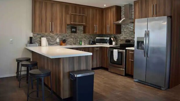 A modern kitchen with wooden cabinets, a stainless steel refrigerator, and a central island with stools. - Cancun Street (Weslaco)