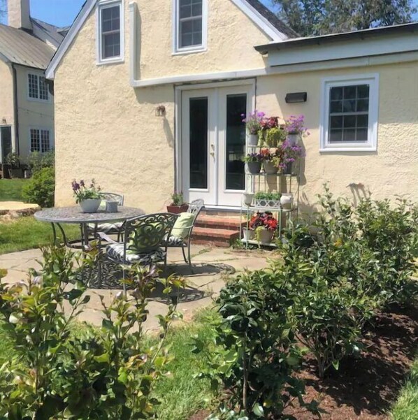 Une maison avec un patio, une table à manger et des chaises. On y trouve des plantes en pot et un petit espace jardin.
