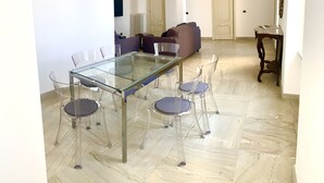 A dining area with a glass-top table and four chairs, a sofa, and a wooden side table in a room with white walls and a marble floor. - Crispi 2enty stylish flat at Spanish Steps (Roma)