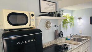 A kitchen with a black Galanz stove, a white microwave, a sink, and a wall-mounted shelf with kitchen items. - Casita Chica (McAllen)