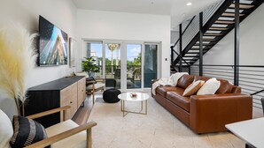 A modern living room with a brown leather sofa, a white coffee table, and a black cabinet. There is a staircase with black railings and a glass door leading outside. - Modern & Gorgeous Rooftop City Views & Close to the Best of Everything in SA! (San Antonio)
