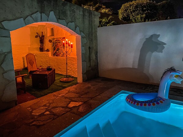 A stone-walled outdoor area with a pool, a wooden chair, a table with a vase, and a wall with a shadow of a person.