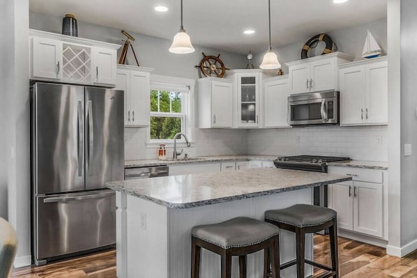 A modern kitchen with white cabinetry, stainless steel appliances, a marble island with stools, and nautical decor.