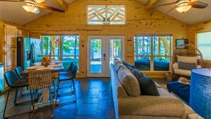 A spacious wooden cabin with a dining area, a sofa, and a large window overlooking a forest.
