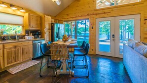 A wooden cabin interior with a dining area, kitchen, and a large window overlooking a forest.
