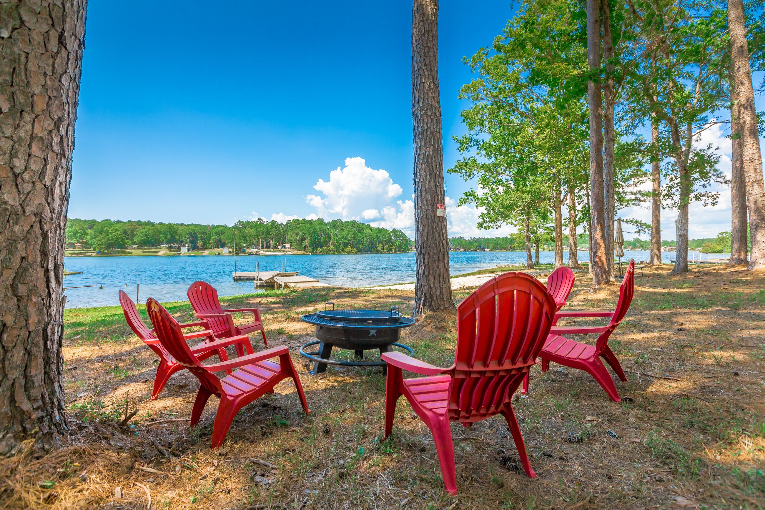 Un cadre idyllique au bord du lac, avec des fauteuils Adirondack rouges, un foyer et une vue sur le lac et les arbres qui s’étendent au loin.