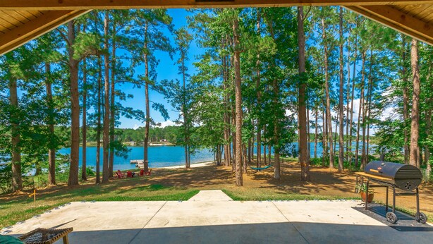 A covered outdoor area with a concrete patio, a hammock, a grill, and a view of a lake surrounded by tall trees.
