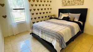 A bedroom with a bed, a window with curtains, a wall with heart patterns, and a floral painting. - Sundown Stay (San Juan)