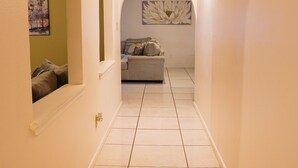 Un couloir étroit, doté d’un sol carrelé et de portes en arc, mène à un espace de vie comprenant un canapé et une œuvre d’art accrochée au mur.