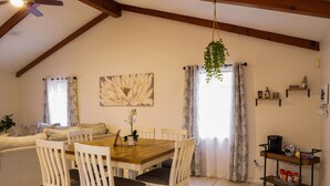 Un coin repas avec une table en bois et des chaises blanches, un ventilateur de plafond, une plante suspendue et un chariot de cuisine avec des bocaux.