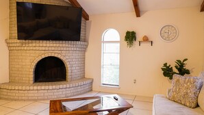 Un salon avec une cheminée en briques, un téléviseur à écran plat fixé au-dessus, un canapé agrémenté de coussins à motifs et une horloge au mur.