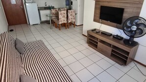 A living room with a sofa, TV, and fan. There is a kitchen in the background with a fridge, microwave, and chairs. - Apartamento Catamarã Portiville (Maceió)
