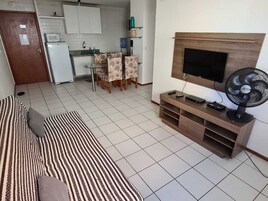 Sala de estar com sofá, TV e ventilador. Cozinha ao fundo, com geladeira, micro-ondas e cadeiras.