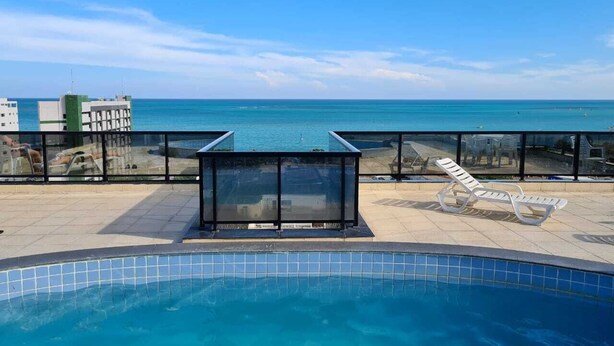 A swimming pool with a glass enclosure and lounge chairs, overlooking a clear blue sea. - Apartamento Catamarã Portiville (Maceió)