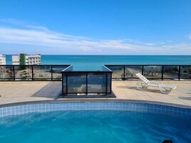 Piscina com vedação de vidro e espreguiçadeiras, com vista para o mar azul e límpido.