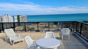 A terrace with a table and chairs, overlooking a body of water and buildings. - Apartamento Catamarã Portiville (Maceió)