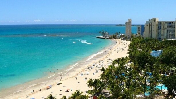 Una playa con aguas turquesas cristalinas, una orilla de arena y una zona turística con palmeras y edificios al fondo.
