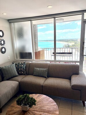 A modern living room with a large window, a sectional sofa, decorative pillows, a round wooden coffee table, and a small potted plant.