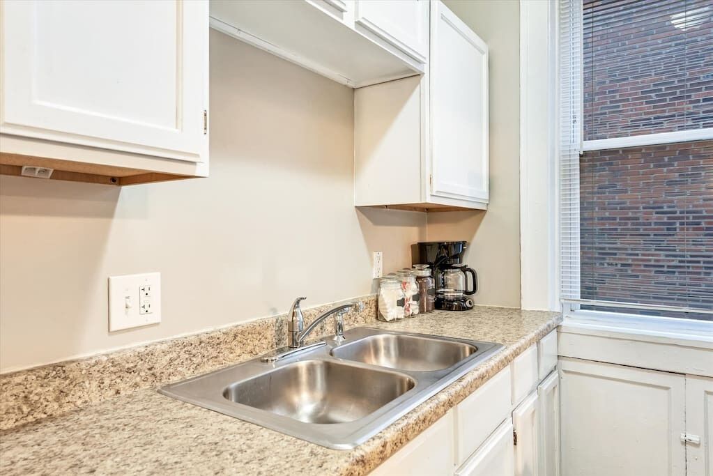A kitchen with two stainless steel sinks, white cabinets, a granite countertop, a coffee maker, and a window with blinds.