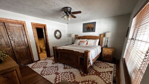 Une chambre à coucher avec un lit en bois, un ventilateur de plafond, une table de chevet, une commode, un tapis et une fenêtre avec des stores.