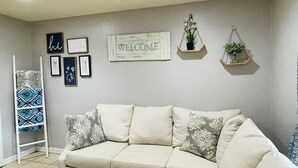 Un salon avec un canapé d’angle beige, une échelle et des éléments de décoration murale, notamment une pancarte « BIENVENUE » et des tableaux encadrés.