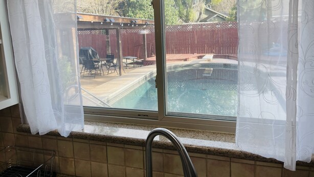 Une cuisine avec une fenêtre donnant sur une piscine située dans le jardin, un espace patio et une clôture en bois.
