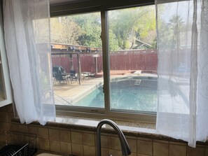 A kitchen with a window overlooking a backyard swimming pool, a patio area, and a wooden fence.