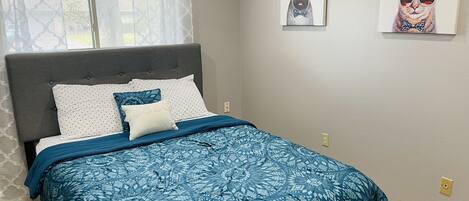 A bedroom with a bed, a grey upholstered headboard, patterned bedding, and two wall-mounted artworks.