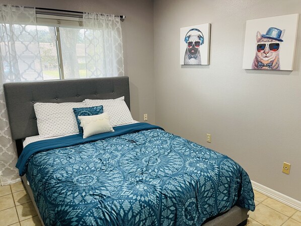 A bedroom with a bed, a grey upholstered headboard, patterned bedding, and two wall-mounted artworks.