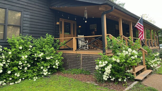 Une maison en bois avec un porche, un drapeau et des arbustes à fleurs blanches.