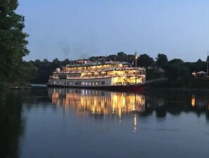 Ein großes, mehrstöckiges Flussboot mit beleuchteten Oberdecks, liegt an einem ruhigen Fluss festgemacht.
