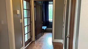 Un pasillo con piso de madera, una puerta que da a una habitación con cama y una ventana con cortinas.