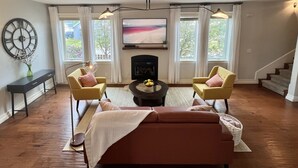 Una sala de estar con un sofá, dos sillones, una mesita de centro, chimenea, televisor y ventanas amplias.