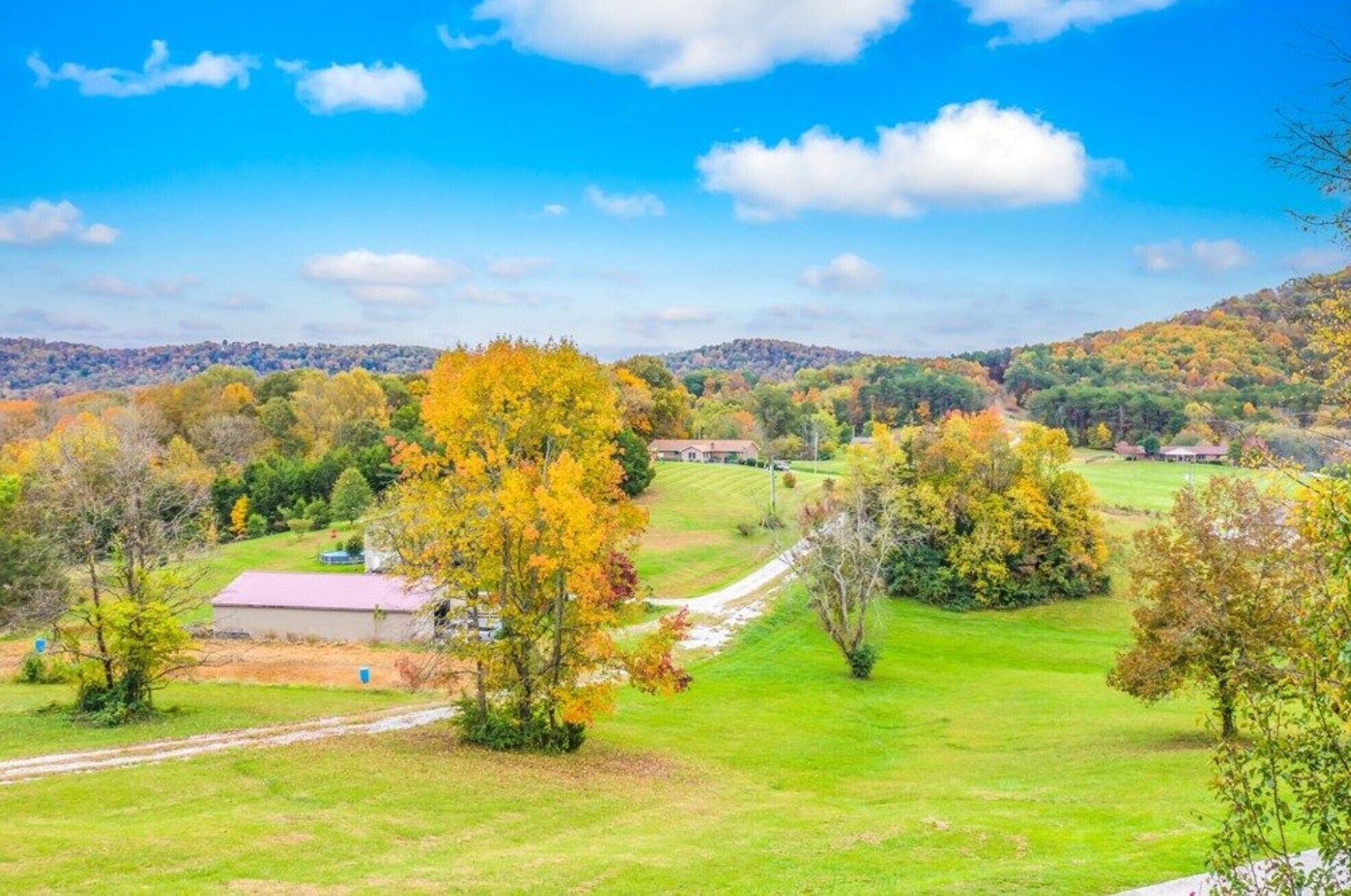 A rural landscape with a house, green fields, and autumn-colored trees.