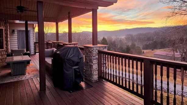 Una terraza de madera con mobiliario exterior y una parrilla, con vistas a un paisaje pintoresco al atardecer.